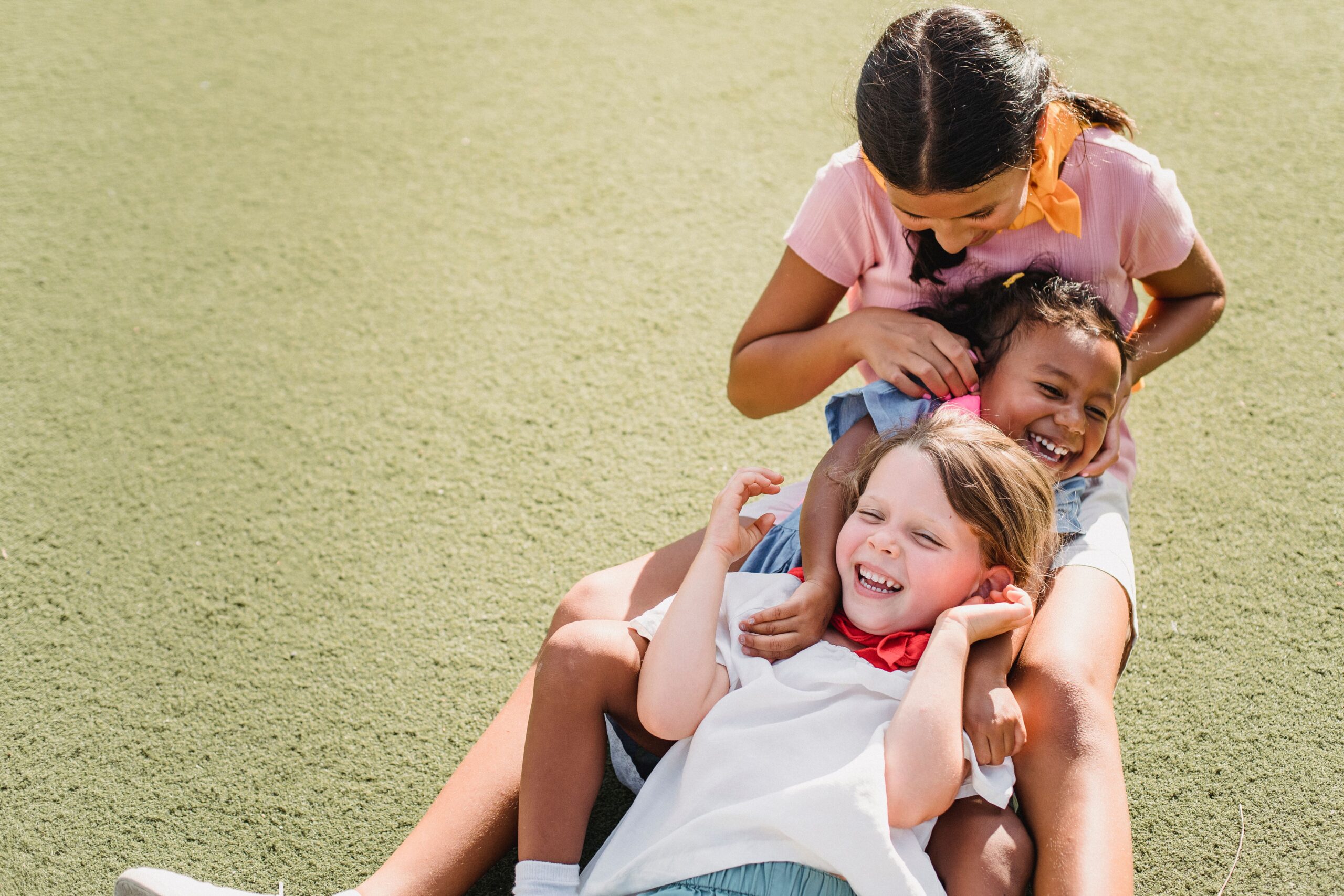 Three children laughing and playing together on grass under the sunny sky, showcasing pure joy and friendship.